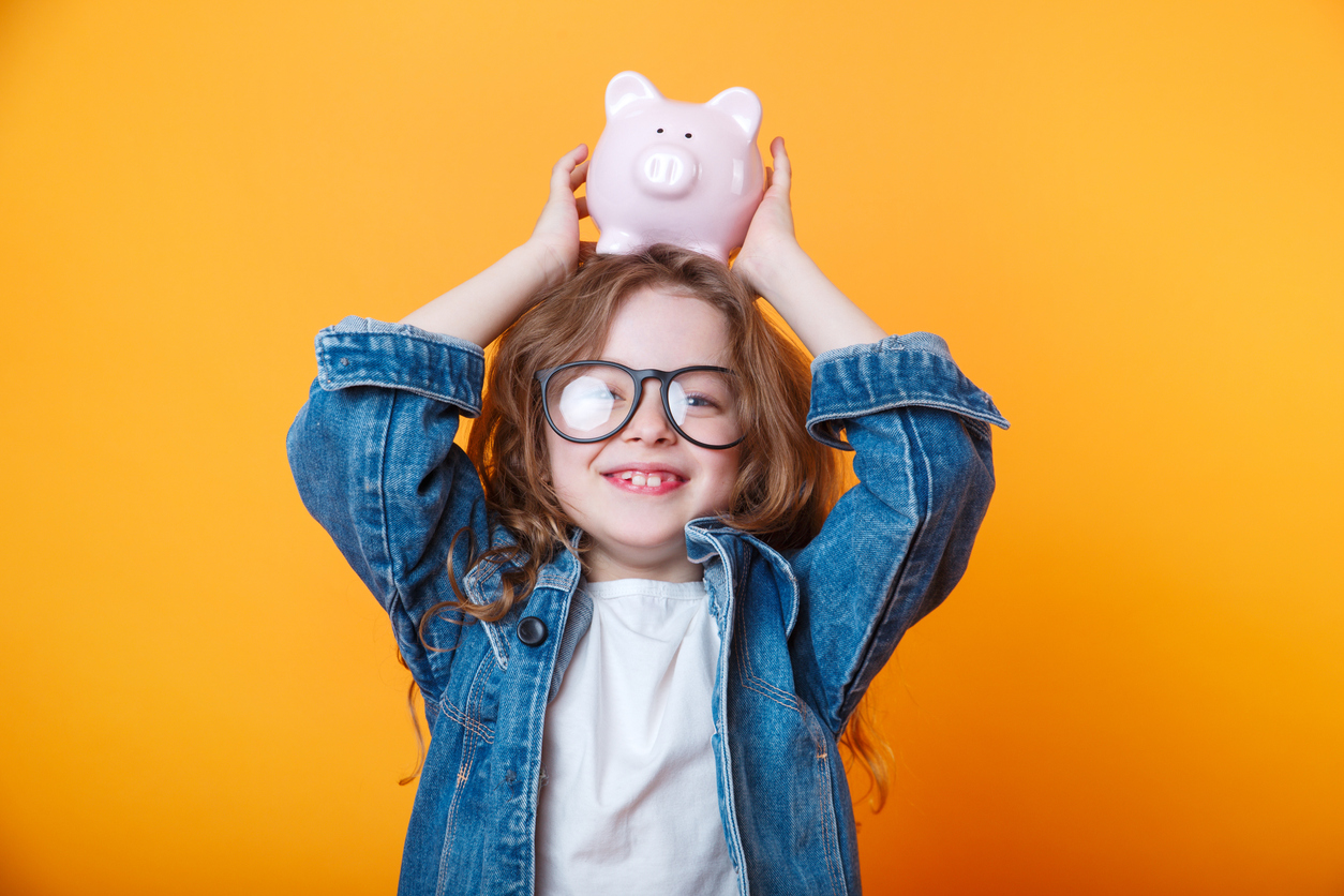 ING Junior Depot: Niedliches kleines Mädchen mit Brille schüttelt Sparschwein auf orangefarbenem Hintergrund.